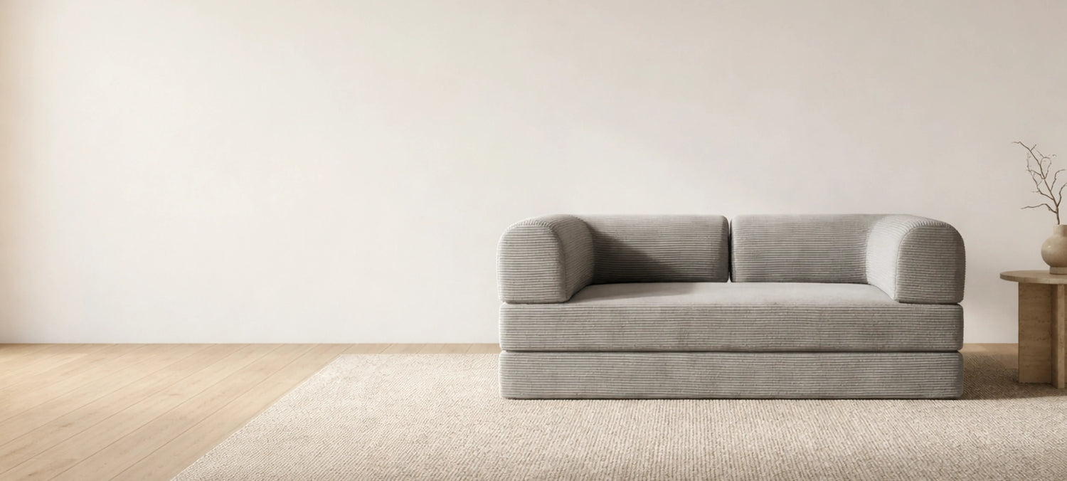 Sala minimalista con sofá gris claro de líneas rectas y redondeadas, alfombra beige y mesa auxiliar con jarrón decorativo.