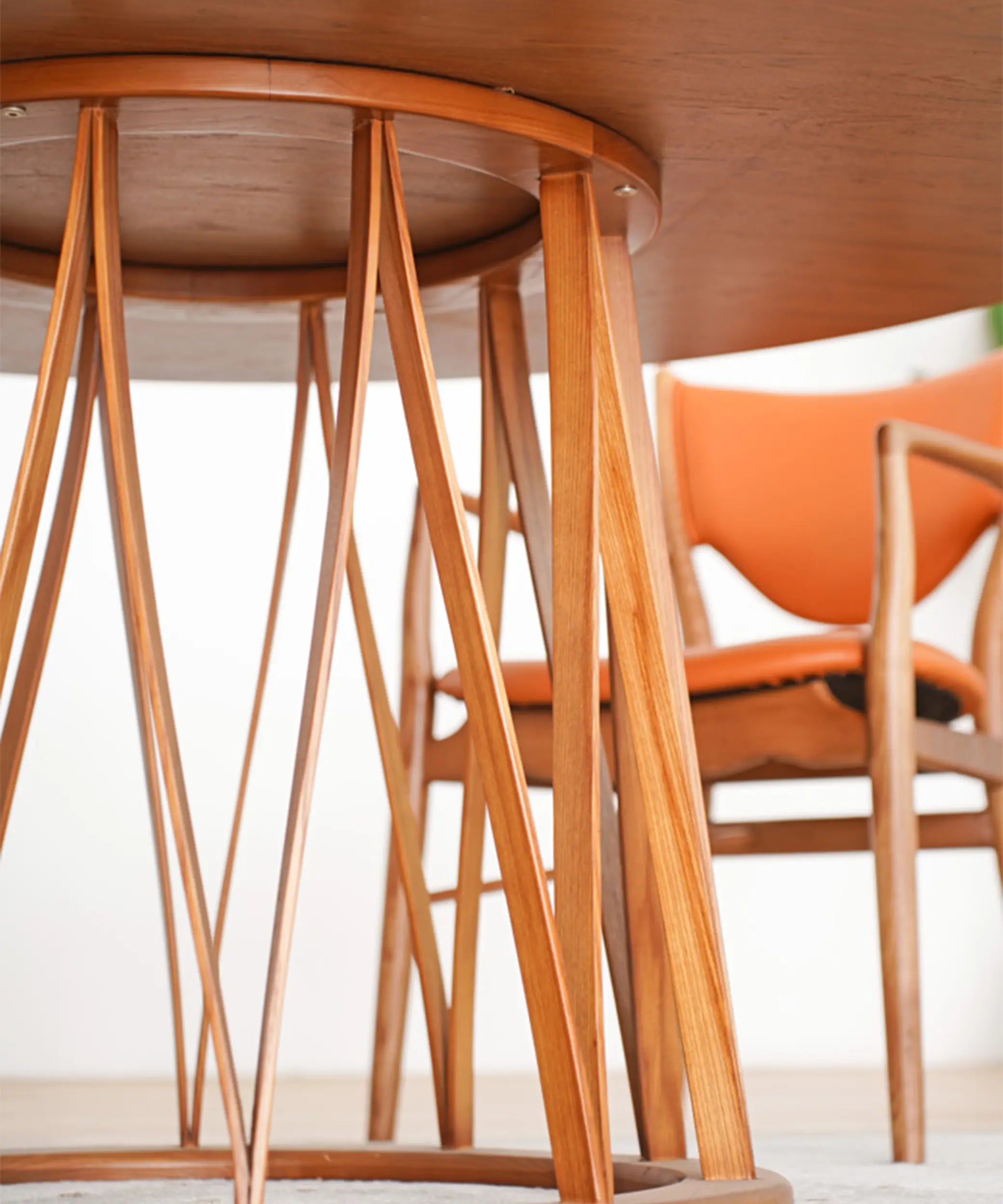 Mesa redonda de madera con patas delgadas entrelazadas y silla con respaldo naranja en fondo claro.
